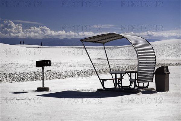 USA, New Mexico, Otero County, White Sands National Monunment. Landscape of white sand with dramatic cloudscape above. Seating area and distant figures. 
Photo : Hugh Rooney
