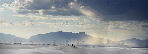 USA, New Mexico, Otero County, White Sands National Monunment. Landscape of white sand with dramatic cloudscape cut through by sunlight above. Group of people in distance. 
Photo : Hugh Rooney