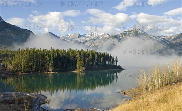 Canada, Alberta, Kananaskis, Early morning mist rising from Barrier Lake with Heart Mountain and the Rockies in the background reflection of pine trees in lake. 
Photo : Trevor Page
