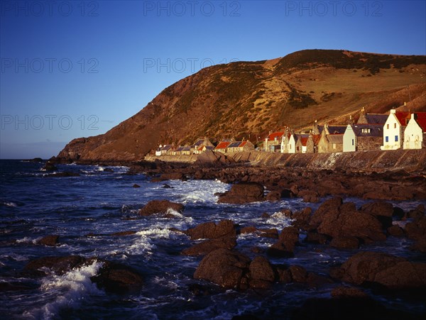 Scotland, Aberdeenshire, Crovie, One time fishing village seen from shoreline. Row of cottages at foot of steep hillside overlooking coast and stone jetty. 
Photo : Bryan Pickering