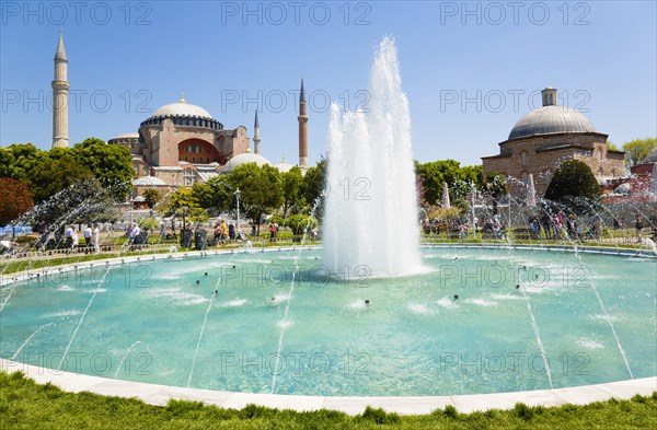 Turkey, Istanbul, Sultanahmet Haghia Sophia with dome and minarets beyond the water fountain and Baths of Roxelana in the gardens with sightseeing tourists. 
Photo : Paul Seheult