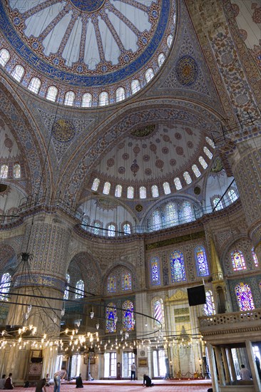 Turkey, Istanbul, Sultanahmet Camii The Blue Mosque interior with people at prayer beneath chandeliers and decorated domes above and stained glass windows beyond. 
Photo : Paul Seheult