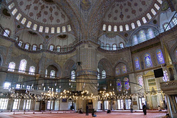 Turkey, Istanbul, Sultanahmet Camii The Blue Mosque interior with people at prayer beneath chandeliers and decorated domes above and stained glass windows beyond. 
Photo : Paul Seheult