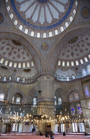 Turkey, Istanbul, Sultanahmet Camii The Blue Mosque interior with people at prayer beneath chandeliers and decorated domes above and stained glass windows beyond. 
Photo : Paul Seheult