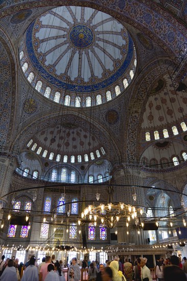 Turkey, Istanbul, Sultanahmet Camii The Blue Mosque interior with sightseeing tourists by a chandelier below the decorated domes with stained glass windows beyond. 
Photo : Paul Seheult