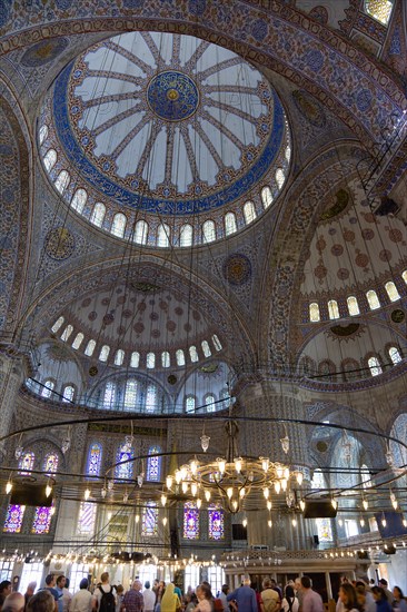 Turkey, Istanbul, Sultanahmet Camii The Blue Mosque interior with sightseeing tourists by a chandelier below the decorated domes with stained glass windows beyond. 
Photo : Paul Seheult