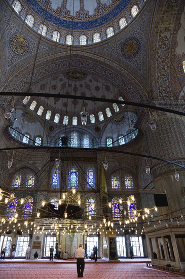 Turkey, Istanbul, Sultanahmet Camii The Blue Mosque interior with people at prayer beneath chandeliers and decorated domes above and stained glass windows beyond. 
Photo : Paul Seheult