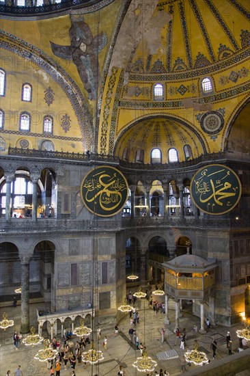 Turkey, Istanbul, Sultanahmet Haghia Sophia Sightseeing tourists beneath the dome with murals and chandeliers in the Nave of the Cathedral with calligraphic roundels of Arabic Koran texts above the Lodge of The Sultan. 
Photo : Paul Seheult