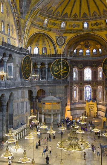 Turkey, Istanbul, Sultanahmet Haghia Sophia Sightseeing tourists beneath the dome with murals and chandeliers in the Nave of the Cathedral with calligraphic roundels of Arabic Koran texts above the Lodge of The Sultan. 
Photo : Paul Seheult