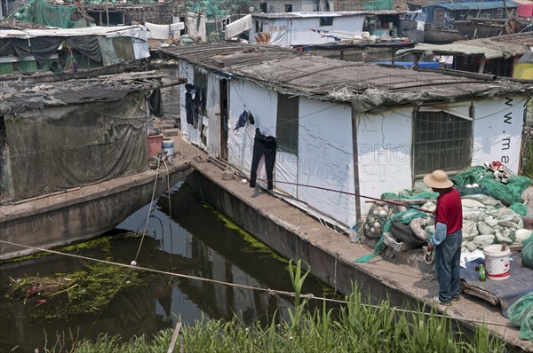 China, Jiangsu, Qidong, Man in a red shirt and straw hat fishing with a rod and line off his boat moored in a canal. Pair of black trousers hanging on a line against a white background; Many poor landless Chinese live their entire lives on boats and make a living hauling goods along inland waterways. 
Photo : Trevor Page