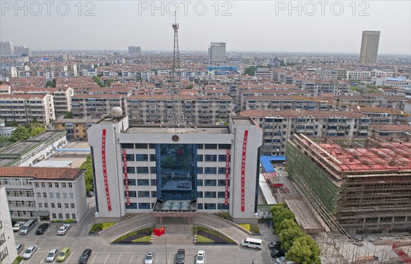 China, Jiangsu, Qidong, Bureau of Public Security with Chinese flag flying in forecourt and radio communications mast on the roof; red banners with Chinese characters hanging from building City skyline and new construction. 
Photo : Trevor Page