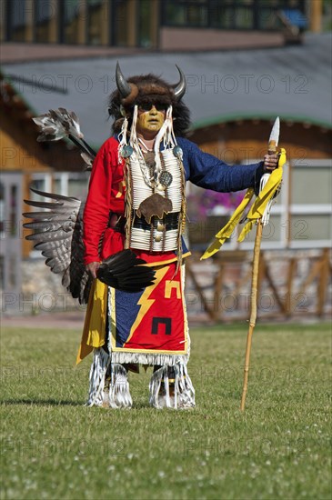 Blackfoot dancer in a buffalo headdress and sunglasses wearing a porcupine quill breastplate and holding a spear and feather fan at the Blackfoot Arts & Heritage Festival Pow Wow organized by Parks Canada and the Blackfoot Canadian Cultural Society at this UNESCO World Heritage Site red blue yellow
