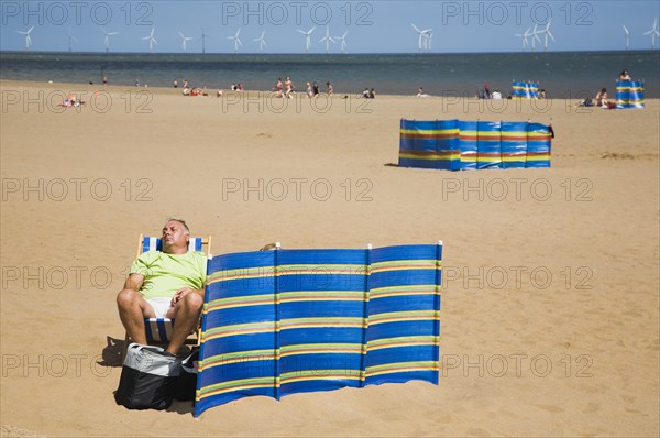 Man sleeps in sunshine behind striped windbreak with Lincs Wind Farm offshore behind.