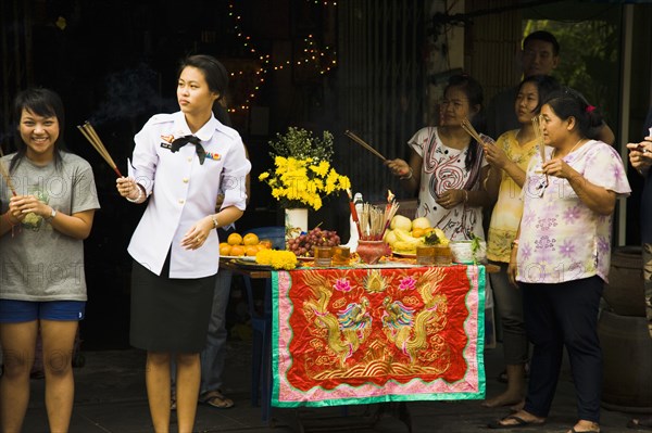 Woman in military uniform holding joss sticks beside offerings on decorated table celebrating local temple parade.