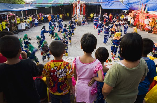 Thai children watch dance troupe at local temple from elevated stage.