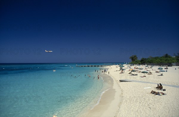 West Indies, Jamaica, Montego Bay, Sandy beach beside aquamarine water with people sunbathing and in the sea with low flying aeroplane over the water. Photo : David Cumming