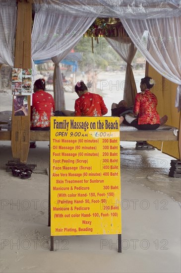 Thailand, Koh Samui , Chaweng Beach, Beach Massage with three women wearing red sat on benches giving massage with a sign displaying prices for various treatments in the foreground. Photo : Derek Cattani