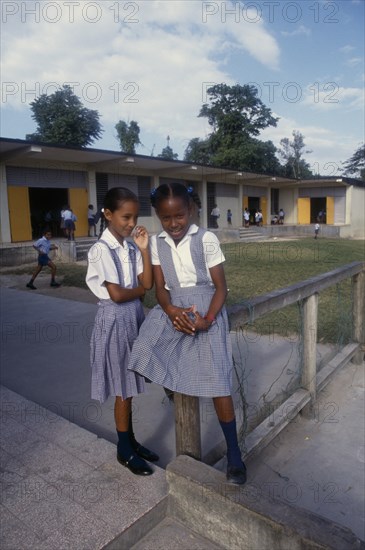 West Indies, Jamaica, Education, Two schoolgirls in checked dresses in playground in front of school buildings. Photo : David Cumming