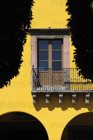 Mexico, Bajio, San Miguel de Allende, El Jardin detail of yellow painted exterior facade of colonial mansion with French windows and balcony part framed by silhouetted trees. Photo : Nick Bonetti