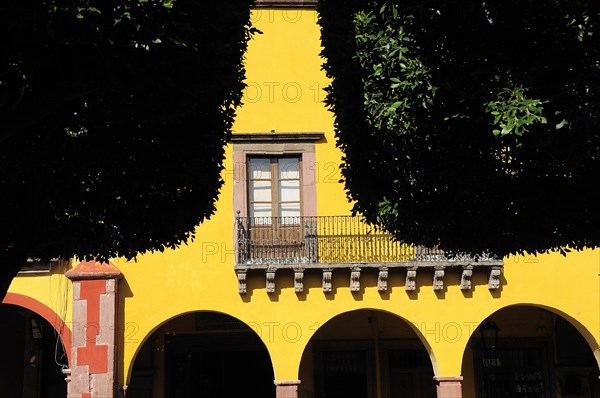 Mexico, Bajio, San Miguel de Allende, El Jardin Detail of yellow painted facade of colonial mansion with French window and balcony part framed by trees in foreground. Photo : Nick Bonetti