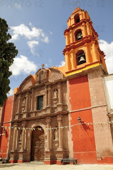 Mexico, Bajio, San Miguel de Allende, Exterior facade and bell tower of orange and yellow painted church building with flags hanging across doorway.. Photo : Nick Bonetti