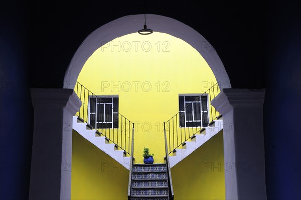 Mexico, Puebla, Looking into inner courtyard through archway in shadow towards yellow painted wall with blue and white tiled steps plant in blue pot and pair of square recessed windows. Photo : Nick Bonetti