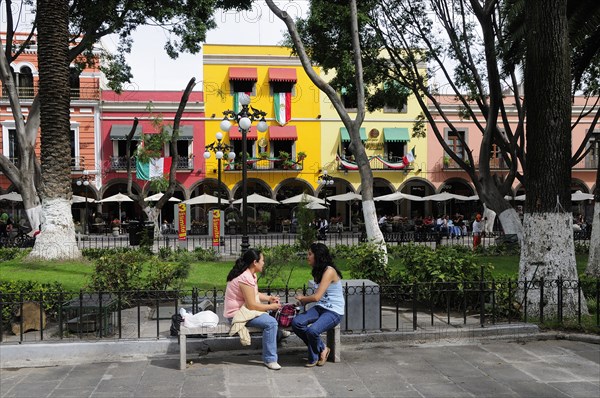 Mexico, Puebla, Two young women chatting on stone bench in the Zocalo with colourful building facades hung with Mexican flags and line of sun umbrellas and outside cafe tables beyond area of grass behind. Photo : Nick Bonetti