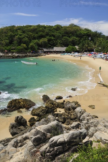 Mexico, Oaxaca, Puerto Escondido, View onto Playa Manzanillo beach with rocks in foreground and tourist boats people and tree covered headland beyond. Photo : Nick Bonetti