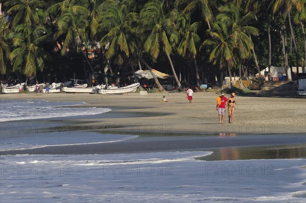 Mexico, Oaxaca, Puerto Escondido, Playa Marinera with couple walking along shore line of boats pulled up onto sand and overhanging palms beyond. Photo : Nick Bonetti