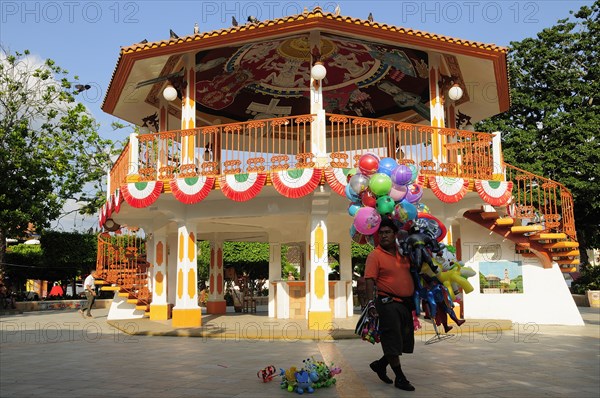 Mexico, Veracruz, Papantla, Balloon seller walking past brightly painted bandstand in the Zocalo. Photo : Nick Bonetti
