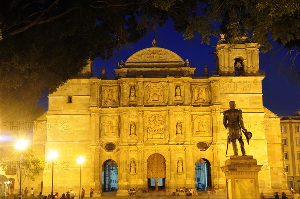 Mexico, Oaxaca, Baroque exterior facade of cathedral at night part framed by tree branches with people and statue in foreground. Photo : Nick Bonetti