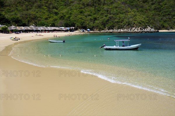 Mexico, Oaxaca, Huatulco, Playa La Entrega Stretch of sandy beach lined with thatched open fronted restaurants with tour boat in shallow water in foreground. Photo : Nick Bonetti