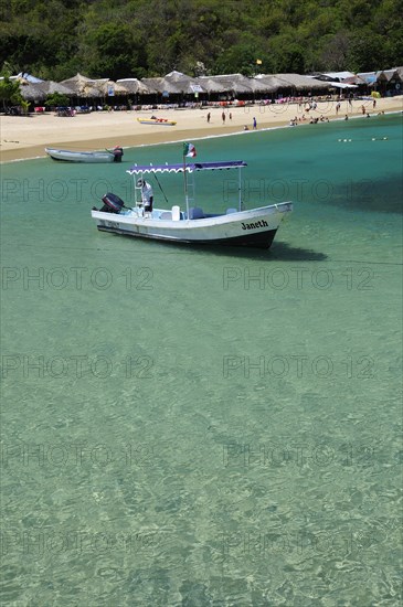 Mexico, Oaxaca, Huatulco, Playa La Entrega Tour boat moored in clear shallow water in foreground of sandy beach lined with thatched open fronted restaurants.. Photo : Nick Bonetti