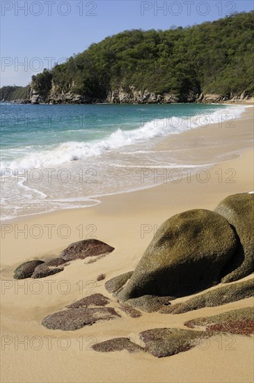 Mexico, Oaxaca, Huatulco, Bahia Chahue Rocks and deserted sandy beach with surf breaking on shore and rocky headland beyond.. Photo : Nick Bonetti