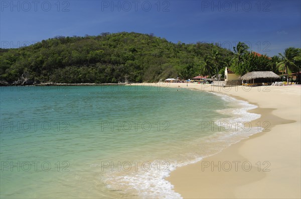 Mexico, Oaxaca, Huatulco, Sandy beach at Bahia Santa Cruz lined by restaurants and other buildings with tree covered coastline beyond. Photo : Nick Bonetti