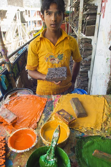 Bangladesh, Dhaka, Young man holding a printing block used to print on cotton fabrics in New Market with pallettes of colours in front of him. 
Photo Nic I Anson / Eye Ubiquitous