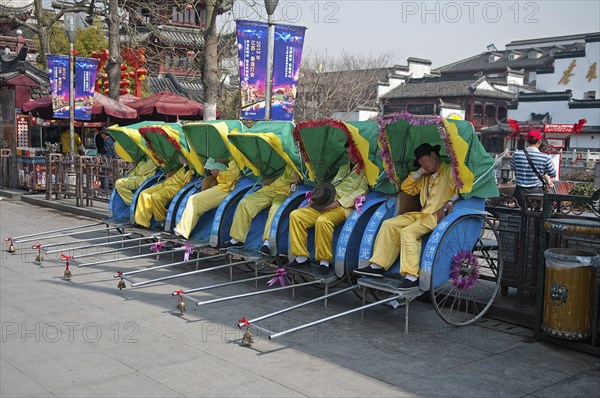 China, Jiangsu, Nanjing, Row of rickshaws with the rickshaw pullers in yellow uniform and hats asleep outside the Fuzi Temple a top tourist spot Fuzi Temple complex tile roofs and red Chinese lanterns in the background. 
Photo Trevor Page / Eye Ubiquitous