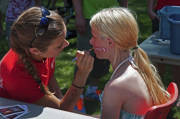 Canada, Alberta, Lethbridge, A bare-shouldered young blonde Canadian girl sits to have the word CANADA painted on her face at Canada Day celebrations at Henderson Park, Face painter in a red T shirt has a maple leaf, Canada's national emblem, stencilled on her forearm. 
Photo Trevor Page