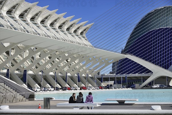 Spain, Valencia Province, Valencia, La Ciudad de las Artes y las Ciencias  City of Arts and Sciences  Principe Felipe Science Museum  General view of the building with girls on seat in the forground and row of Mercedes cars below the building.