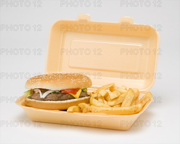 Food, Cooked, Meat, Cheesburger with salad and tomato ketchup in a bun with potato chips inside a polystyrene foam box on a white background.