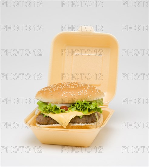 Food, Cooked, Meat, cheesburger with salad and tomato ketchup in a bun inside a polystyrene foam box on a white background.